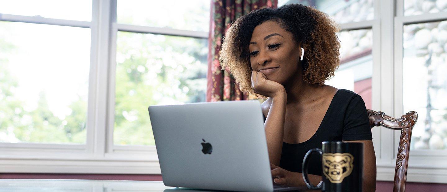 A female student seated at a table with an open laptop and a coffee mug with a gold Oakland University Grizz bear image on it.